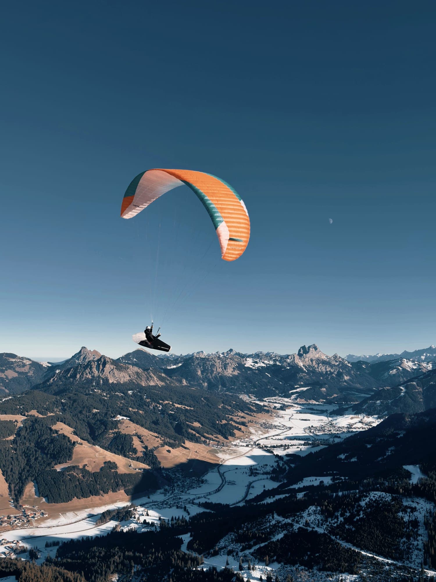 Gleitschirm hoch über einem weiten, teils verschneiten Alpental mit gestaffelten Bergketten im Hintergrund