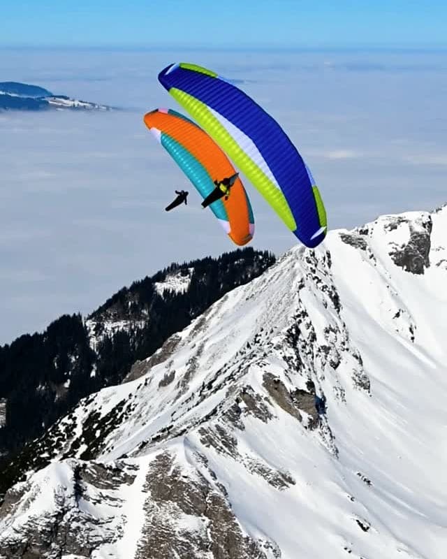 Bunter Gleitschirm über einem scharfen Schneegrat, darunter ein geschlossenes Wolkenmeer